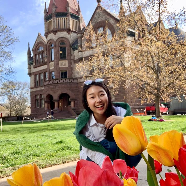 Ga Vin smiling behind red and yellow tulips and standing in front of the Victoria College building
